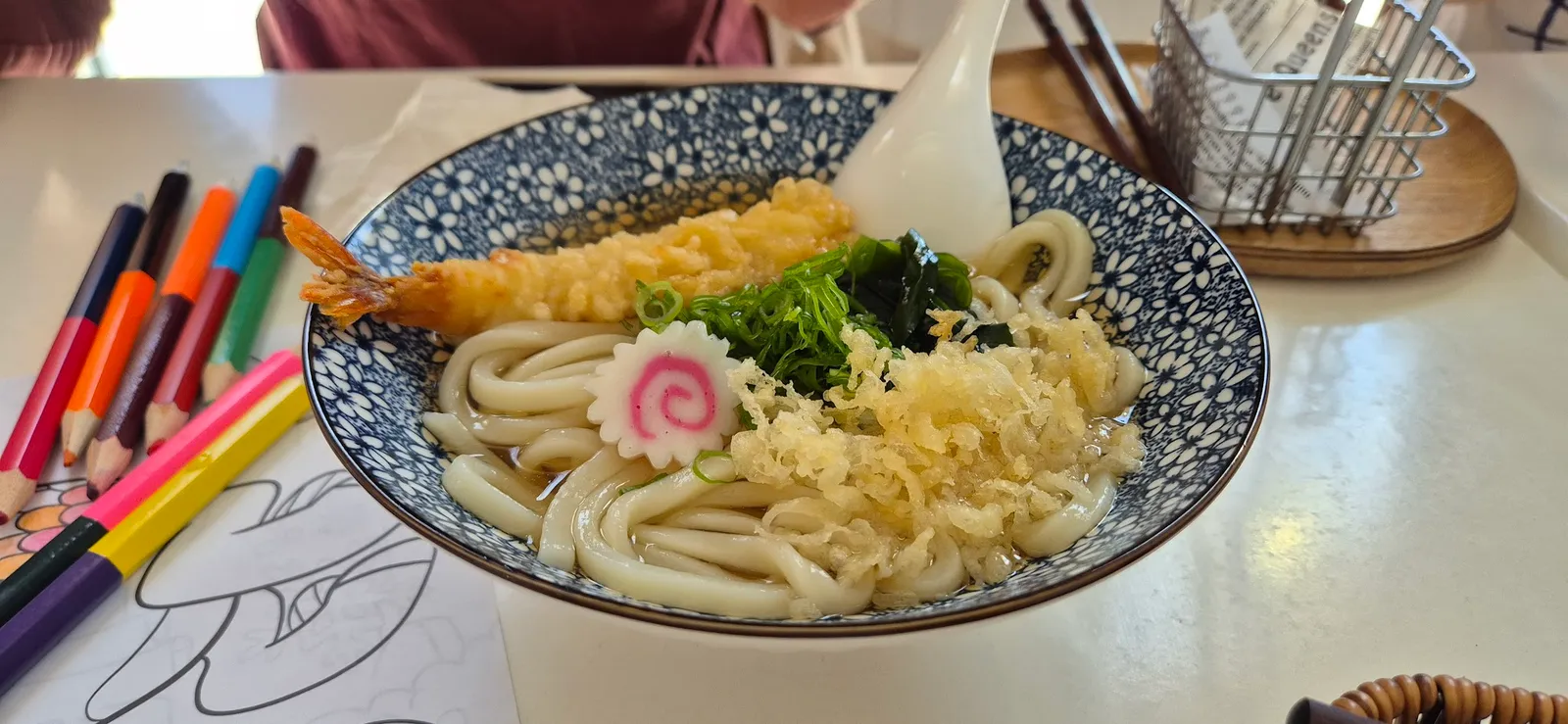 Tempura prawn udon in floral blue bowl, children's crayons on table beside it