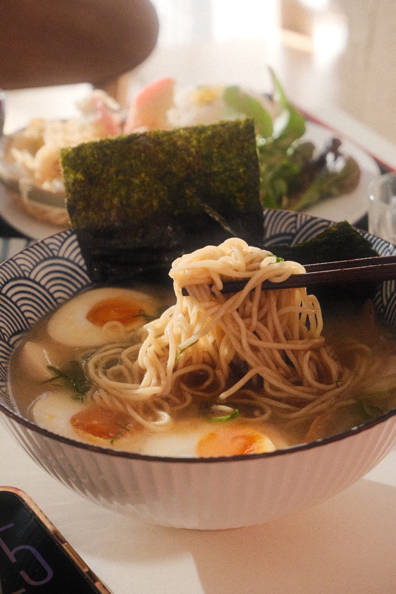 Ramen noodles lifted by chopsticks above broth with egg and nori